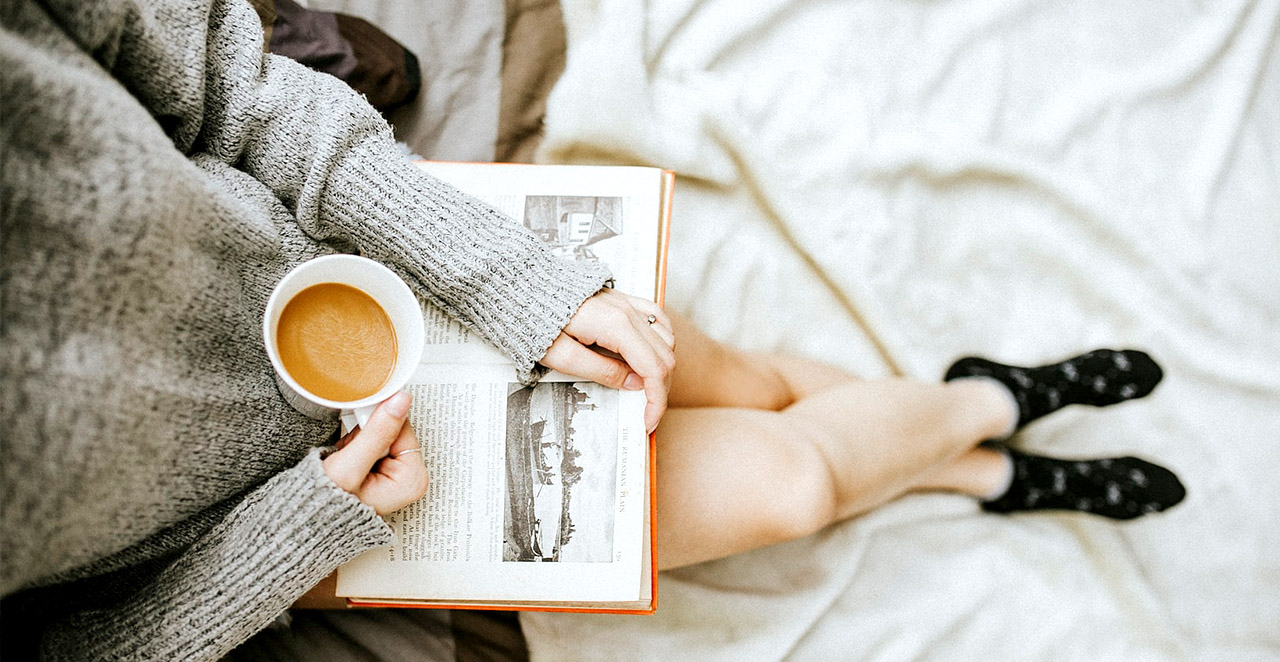 Nutze die Winterzeit zum entspannen. Auf dem Bild sieht man eine Person, die Kafee trinkt und ein Buch liest.