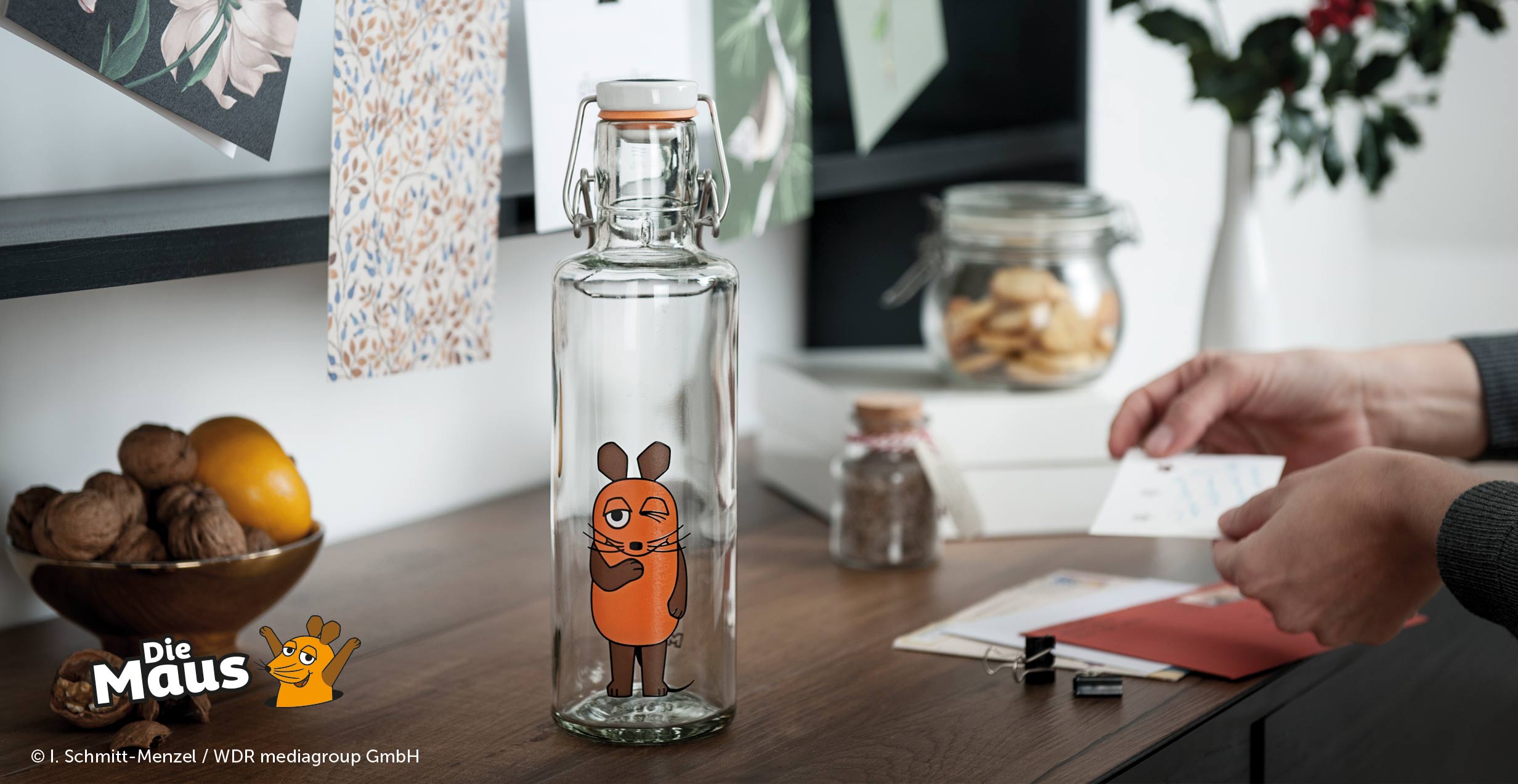 Hier ist eigentlich ein Bild zu sehen. EIne nachhaltige Trinkflasche aus Glas steht auf einem Tisch und auf ihr die Maus abgebildet.